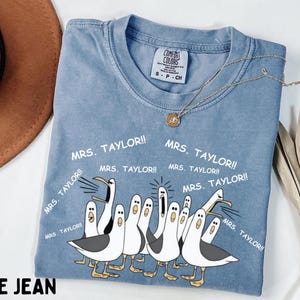 May include: Blue jean colored t-shirt with a graphic of cartoon seagulls and the text "MRS. TAYLOR!!". The shirt has a crew neck and short sleeves. A gold necklace with a pendant is on the shirt.