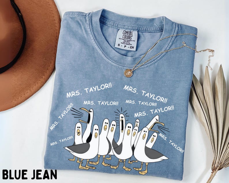 May include: Blue jean colored t-shirt with a graphic of cartoon seagulls and the text "MRS. TAYLOR!!". The shirt has a crew neck and short sleeves. A gold necklace with a pendant is on the shirt.