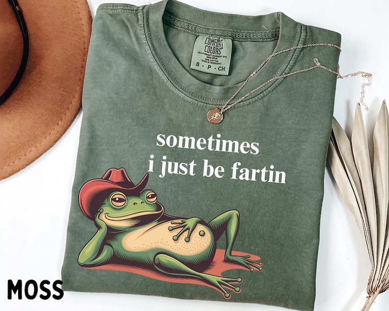 May include: A moss green t-shirt featuring a graphic of a green frog wearing a red cowboy hat and reclining. The text "sometimes i just be fartin" is printed in white above the frog. The shirt has a Comfort Colors brand label at the neck.