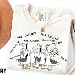May include: Ivory-colored t-shirt featuring a cartoon illustration of seagulls yelling "MRS. TAYLOR!!". The shirt has a crew neck and short sleeves. A gold necklace with a pendant is draped on the shirt. The shirt is labeled "COMFORT COLORS".