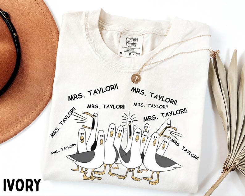 May include: Ivory-colored t-shirt featuring a cartoon illustration of seagulls yelling "MRS. TAYLOR!!". The shirt has a crew neck and short sleeves. A gold necklace with a pendant is draped on the shirt. The shirt is labeled "COMFORT COLORS".