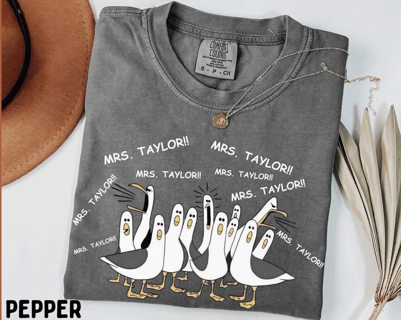 Puede incluir: Camiseta Comfort Colors color pimienta con cuello redondo y mangas cortas. La camiseta presenta un diseño de gaviota de dibujos animados con el texto "MRS. TAYLOR!!". Un collar dorado con un colgante está sobre la camiseta.