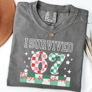 May include: A gray t-shirt with the text "I Survived 67 Days of School" in white. The numbers 6 and 7 are in a checkered pattern of red and green. A gold necklace and a brown hat are also visible.