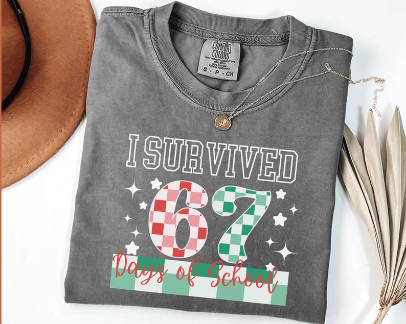 May include: A gray t-shirt with the text "I Survived 67 Days of School" in white. The numbers 6 and 7 are in a checkered pattern of red and green. A gold necklace and a brown hat are also visible.