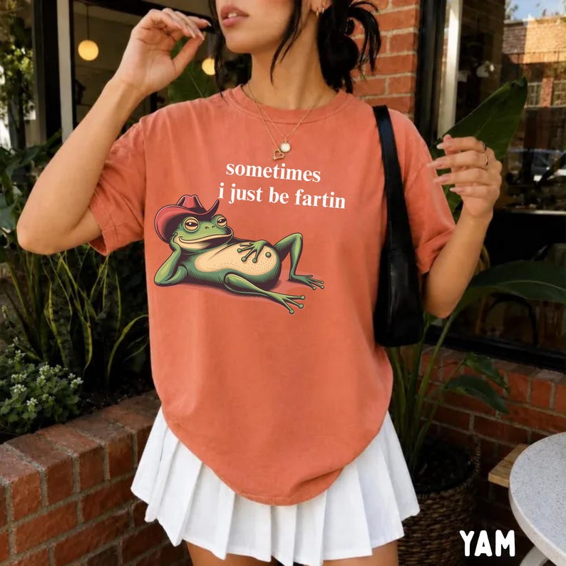 May include: A terracotta-colored short-sleeve t-shirt featuring a graphic of a green frog wearing a red cowboy hat. The text "sometimes i just be fartin" is printed in white above the frog. The shirt is paired with a white pleated skirt.