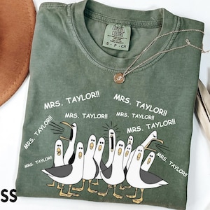 May include: A moss green Comfort Colors t-shirt with a graphic of cartoon seagulls and the text "MRS. TAYLOR!!". The shirt has a crew neck and short sleeves. A gold necklace is draped on the shirt.