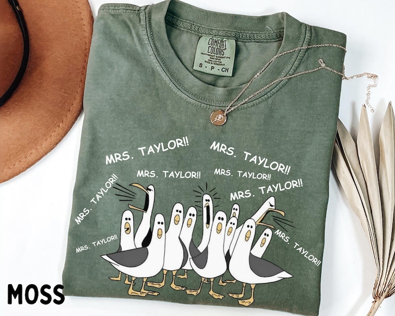 May include: A moss green Comfort Colors t-shirt with a graphic of cartoon seagulls and the text "MRS. TAYLOR!!". The shirt has a crew neck and short sleeves. A gold necklace is draped on the shirt.
