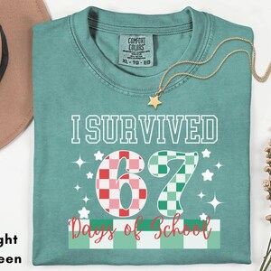 May include: Light green t-shirt with the text "I Survived 67 Days of School". The number 67 is designed with a checkered pattern in red and green. A gold star necklace is draped across the shirt.