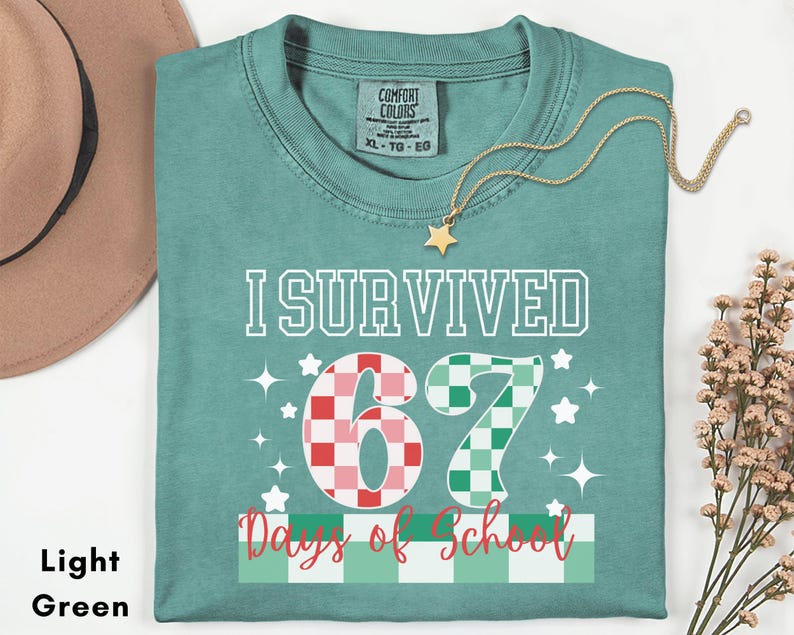 May include: Light green t-shirt with the text "I Survived 67 Days of School". The number 67 is designed with a checkered pattern in red and green. A gold star necklace is draped across the shirt.
