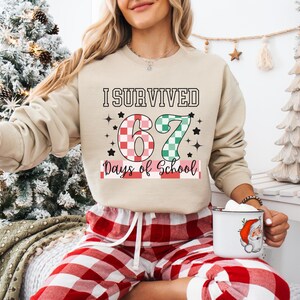 May include: Beige sweatshirt with the text "I Survived 67 Days of School" in black, red, and green. The number 6 is red and white checkered, and the number 7 is green and white checkered. The person is wearing red and white plaid pajama pants.