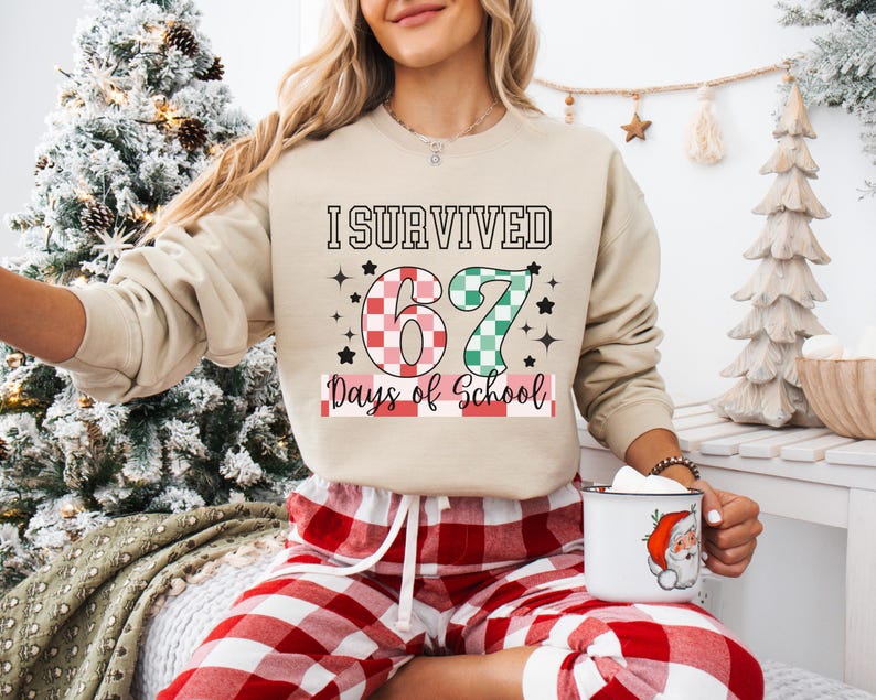 May include: Beige sweatshirt with the text "I Survived 67 Days of School" in black, red, and green. The number 6 is red and white checkered, and the number 7 is green and white checkered. The person is wearing red and white plaid pajama pants.