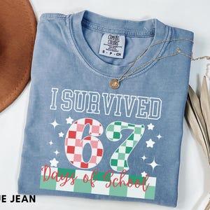 May include: A blue jean Comfort Colors t-shirt with the text "I Survived 67 Days of School." The numbers 6 and 7 are designed with a checkered pattern in red and green. The shirt has a crew neck and short sleeves.