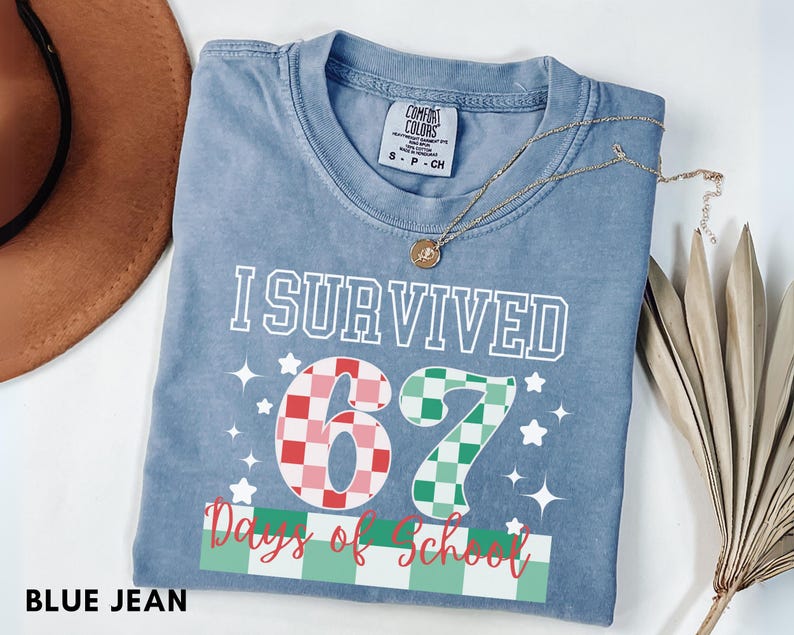 May include: A blue jean Comfort Colors t-shirt with the text "I Survived 67 Days of School." The numbers 6 and 7 are designed with a checkered pattern in red and green. The shirt has a crew neck and short sleeves.