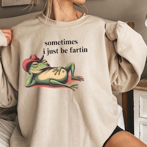 May include: A beige crewneck sweatshirt featuring a graphic of a green frog wearing a red cowboy hat, lying down with its hand on its belly. Above the frog, the black text reads: sometimes i just be fartin. The sweatshirt has long sleeves.