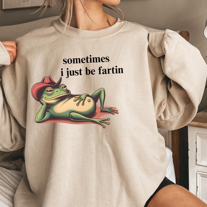 May include: A beige crewneck sweatshirt featuring a graphic of a green frog wearing a red cowboy hat, lying down with its hand on its belly. Above the frog, the black text reads: sometimes i just be fartin. The sweatshirt has long sleeves.