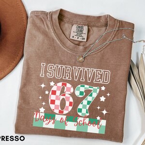 May include: Espresso-colored t-shirt with the text "I Survived 67 Days of School." The number "67" is designed with a checkered pattern in red, pink, and green. The shirt has a crew neck and short sleeves. A silver necklace with a pendant is on the shirt.