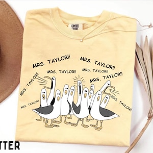 May include: Butter-colored t-shirt with a cartoon illustration of seagulls yelling "MRS. TAYLOR!!" in black text. The shirt has a crew neck and short sleeves. A gold necklace is draped on the shirt.