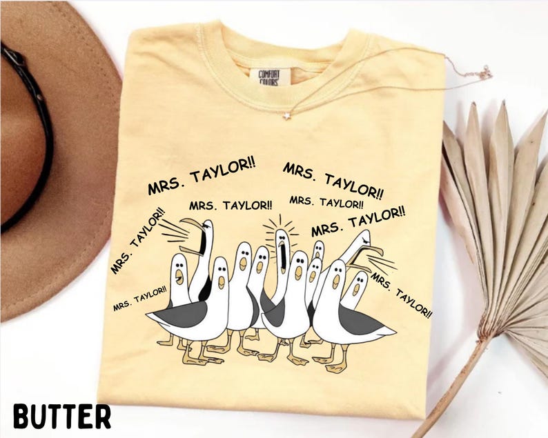 May include: Butter-colored t-shirt with a cartoon illustration of seagulls yelling "MRS. TAYLOR!!" in black text. The shirt has a crew neck and short sleeves. A gold necklace is draped on the shirt.
