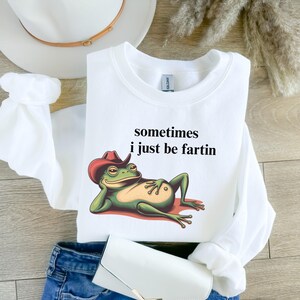 May include: A white long-sleeve sweatshirt featuring a graphic of a green frog wearing a red cowboy hat, reclining on a red surface. The text above the frog reads, sometimes i just be fartin. The sweatshirt has a crew neckline and ribbed cuffs.