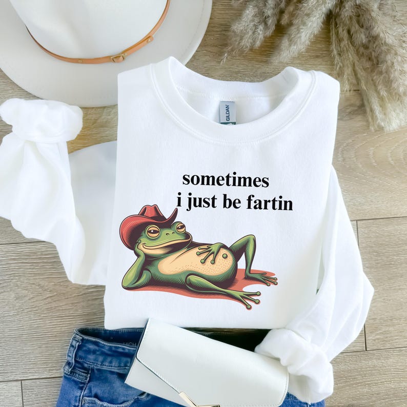 May include: A white long-sleeve sweatshirt featuring a graphic of a green frog wearing a red cowboy hat, reclining on a red surface. The text above the frog reads, sometimes i just be fartin. The sweatshirt has a crew neckline and ribbed cuffs.