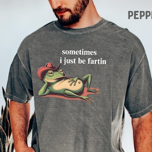 May include: A charcoal gray short-sleeve t-shirt featuring a graphic of a green frog wearing a red cowboy hat, reclining on its side. The text above the frog reads, sometimes i just be fartin. The shirt has a washed, vintage-style texture.