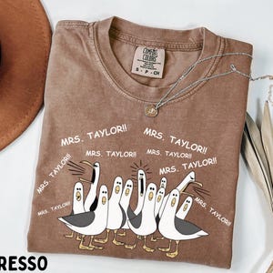 May include: Espresso-colored t-shirt featuring a cartoon illustration of seagulls with the text "MRS. TAYLOR!!" repeated around the image. The shirt has a crew neck and a tag that reads "COMFORT COLORS". A silver necklace with a pendant is draped across the shirt.