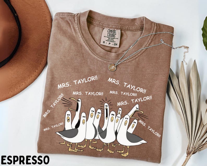 May include: Espresso-colored t-shirt featuring a cartoon illustration of seagulls with the text "MRS. TAYLOR!!" repeated around the image. The shirt has a crew neck and a tag that reads "COMFORT COLORS". A silver necklace with a pendant is draped across the shirt.