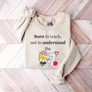 May include: Cream-colored sweatshirt with the text "Born to teach, not to understand the 67". The number 67 is designed with a pencil, book, and apple. The sweatshirt is folded on a wooden surface.