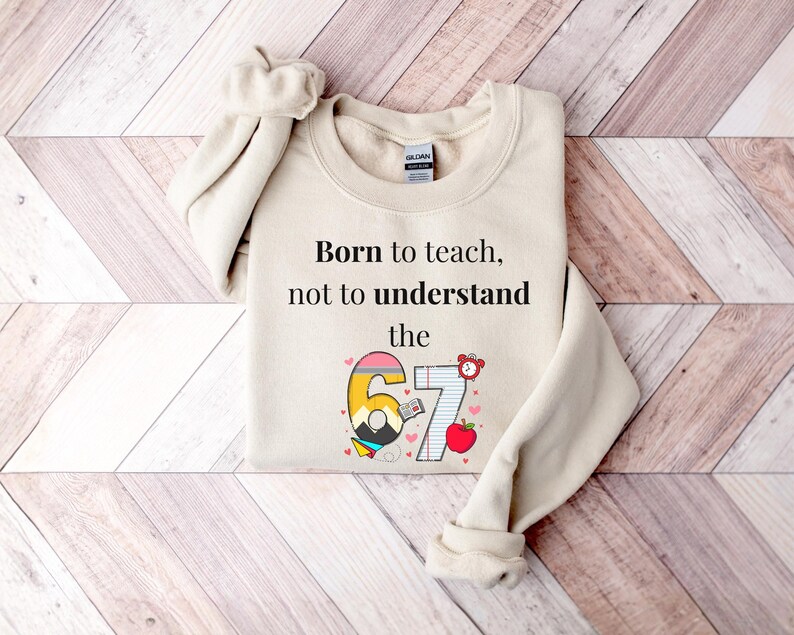 May include: Cream-colored sweatshirt with the text "Born to teach, not to understand the 67". The number 67 is designed with a pencil, book, and apple. The sweatshirt is folded on a wooden surface.