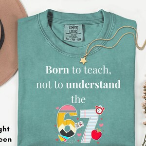 May include: Light green t-shirt with the text "Born to teach, not to understand the 67". The graphic includes a number 6 and 7 with school-themed illustrations. A gold star necklace and a brown hat are also visible.