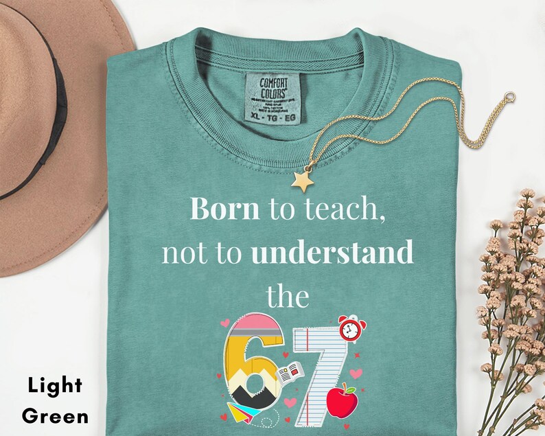 May include: Light green t-shirt with the text "Born to teach, not to understand the 67". The graphic includes a number 6 and 7 with school-themed illustrations. A gold star necklace and a brown hat are also visible.