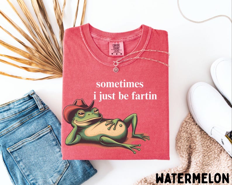 May include: A watermelon-colored t-shirt featuring a graphic of a green frog wearing a brown cowboy hat, reclining while resting its head on one hand. The text "sometimes i just be fartin" is printed in white above the frog illustration.