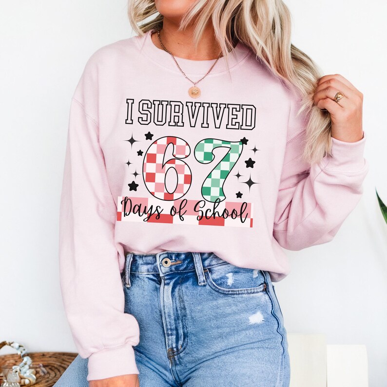 May include: A light pink sweatshirt with the text "I Survived 67 Days of School." The number 67 is designed with a red and white checkered pattern and a green and white checkered pattern. Black stars are scattered around the text.
