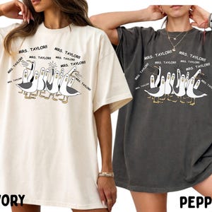 May include: Ivory and pepper-colored oversized t-shirts. Each shirt features a cartoon illustration of seagulls with the text "MRS. TAYLOR!!" repeated around them. The ivory shirt has "IVORY" printed at the bottom, and the pepper shirt has "PEPPER".