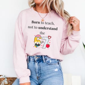 May include: A light pink sweatshirt with the text "Born to teach, not to understand the 67". The graphic includes a colorful number 6 and 7, a red apple, and a small alarm clock. The sweatshirt is paired with blue jeans.