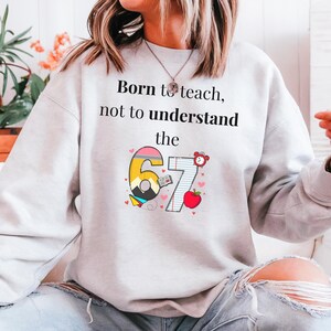 May include: A light gray sweatshirt with the text "Born to teach, not to understand the" above a graphic of the numbers "67" with school-themed illustrations. The "6" is yellow, pink, and blue, and the "7" is on lined paper.
