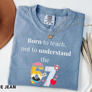 May include: Blue jean colored t-shirt with the text "Born to teach, not to understand the 67". The graphic includes a pencil, apple, and alarm clock. A gold necklace is draped across the shirt.