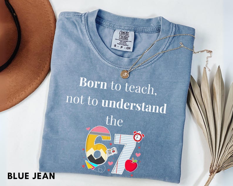 May include: Blue jean colored t-shirt with the text "Born to teach, not to understand the 67". The graphic includes a pencil, apple, and alarm clock. A gold necklace is draped across the shirt.