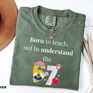 May include: Moss green t-shirt with the text "Born to teach, not to understand the 67". The graphic includes a pencil, apple, and clock. A gold necklace rests on the shirt. A brown hat is in the background.