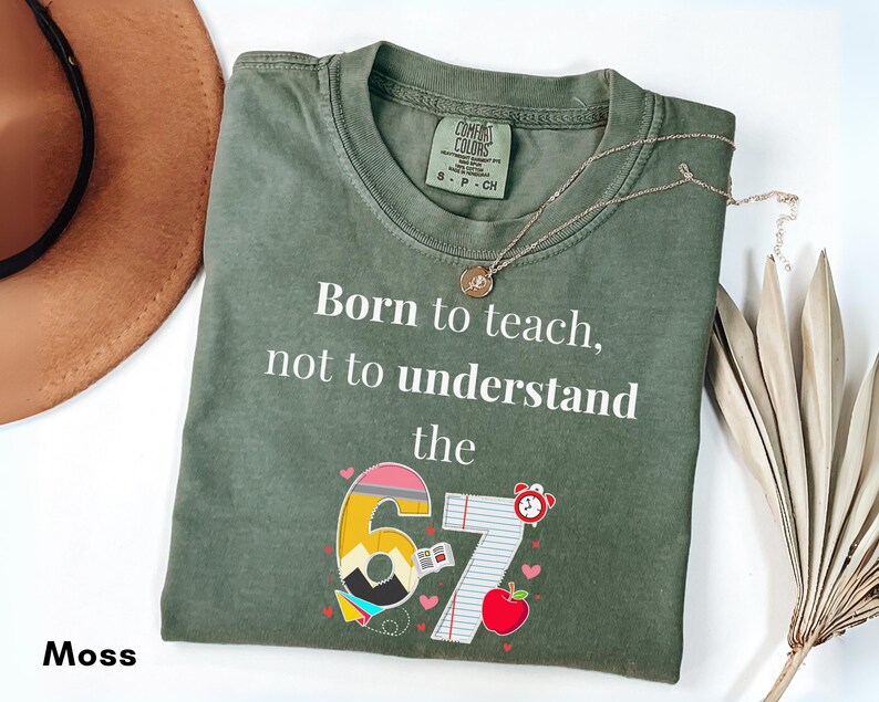 May include: Moss green t-shirt with the text "Born to teach, not to understand the 67". The graphic includes a pencil, apple, and clock. A gold necklace rests on the shirt. A brown hat is in the background.