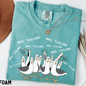 May include: Seafoam green t-shirt with a cartoon illustration of seagulls and the text "MRS. TAYLOR!!" repeated around the birds. The shirt has a crew neck and short sleeves. A gold necklace is draped across the shirt.