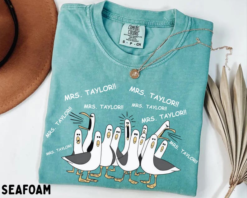 May include: Seafoam green t-shirt with a cartoon illustration of seagulls and the text "MRS. TAYLOR!!" repeated around the birds. The shirt has a crew neck and short sleeves. A gold necklace is draped across the shirt.