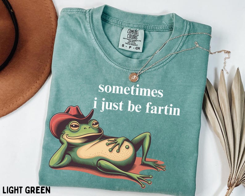 May include: A light green t-shirt featuring a graphic of a green frog wearing a red cowboy hat and reclining. The text "sometimes i just be fartin" is printed in white above the frog. The shirt has a Comfort Colors brand label at the neck.