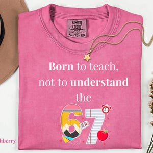 May include: A pink Comfort Colors t-shirt with the text "Born to teach, not to understand the 67." The design includes a pencil, notebook paper, an apple, and a clock. A gold star necklace is draped across the shirt.