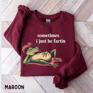 May include: A maroon long-sleeve sweatshirt featuring a graphic of a green frog wearing a red cowboy hat, lying down with one hand behind its head. Above the frog, the white text reads: sometimes i just be fartin.