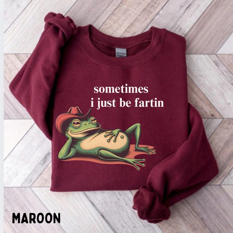 May include: A maroon long-sleeve sweatshirt featuring a graphic of a green frog wearing a red cowboy hat, lying down with one hand behind its head. Above the frog, the white text reads: sometimes i just be fartin.