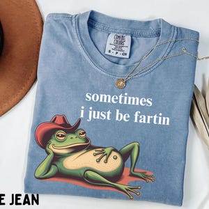 May include: A blue jean colored t-shirt featuring a graphic of a green frog wearing a red cowboy hat, reclining on a red surface. The text "sometimes i just be fartin" is printed in white above the frog. The shirt has a Comfort Colors label.