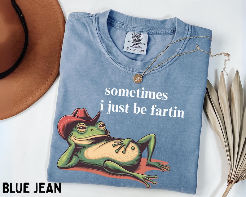 May include: A blue jean colored t-shirt featuring a graphic of a green frog wearing a red cowboy hat, reclining on a red surface. The text "sometimes i just be fartin" is printed in white above the frog. The shirt has a Comfort Colors label.
