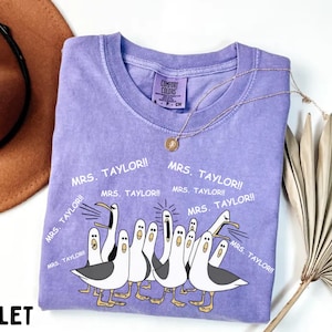 May include: A violet Comfort Colors t-shirt featuring a graphic of seagulls with the text "MRS. TAYLOR!!" repeated around the design. A gold necklace and a brown hat are visible in the background. The word "VIOLET" is printed at the bottom.
