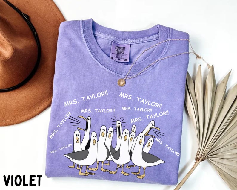 May include: A violet Comfort Colors t-shirt featuring a graphic of seagulls with the text "MRS. TAYLOR!!" repeated around the design. A gold necklace and a brown hat are visible in the background. The word "VIOLET" is printed at the bottom.